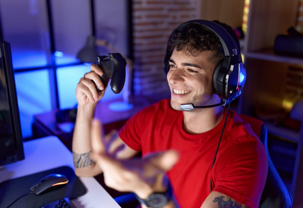 Person mit Headset am Schreibtisch im farbig beleuchteten Streaming-Raum, Controller in der Hand